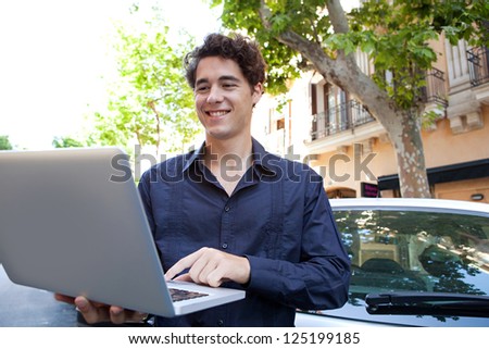Portrait of a stylish businessman standing near a car, using a laptop computer outdoors in the city.