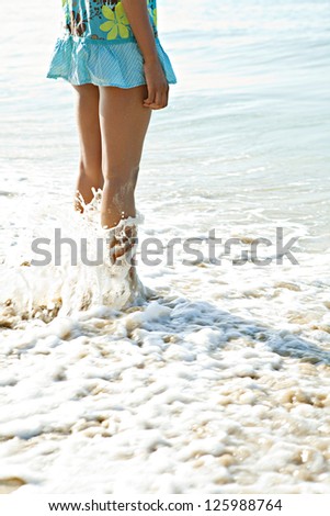 Rear side view of a young woman body on vacations, standing on a white beach shore with her feet in the water, relaxing.