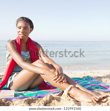 Young attractive black woman sitting on a towel in a white sand beach, relaxing while on vacation and smiling on a sunny day.