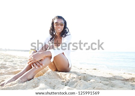 Young attractive black woman sitting on a white sand beach, relaxing while on vacation and smiling on a sunny day.