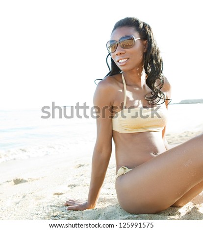 Beautiful young african american woman sitting on a white sand beach near the sea, smiling and wearing a glossy golden bikini and shades.