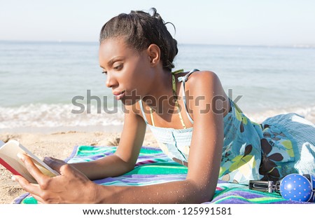 Young attractive african american woman reading a book while laying down on a beach on vacation.