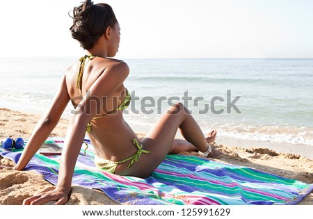 Attractive african american woman on vacation sitting on a towel on a golden beach, contemplating the sea while on vacation, with a blue sky.