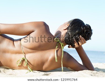Rear close up view of a sexy black woman laying on a white sand beach in a bikini, contemplating the sea on a sunny day on vacations.