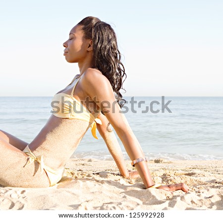 Side portrait of a beautiful black woman sitting on a white sand beach wearing a golden bikini against a blue sky.