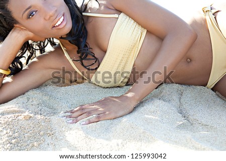 Luxurious perfect beautiful black woman laying down on a white sand beach wearing a golden bikini with shiny beads, relaxing on vacation.