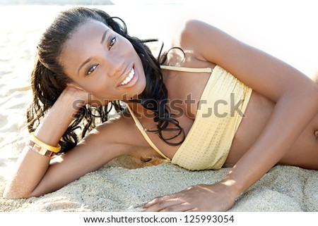 Luxurious perfect beautiful black woman laying down on a white sand beach wearing a golden bikini with shiny beads, relaxing on vacation.
