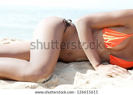 Middle section view of a sexy woman body laying down on a white sand beach, relaxing while on vacation against a blue sea.