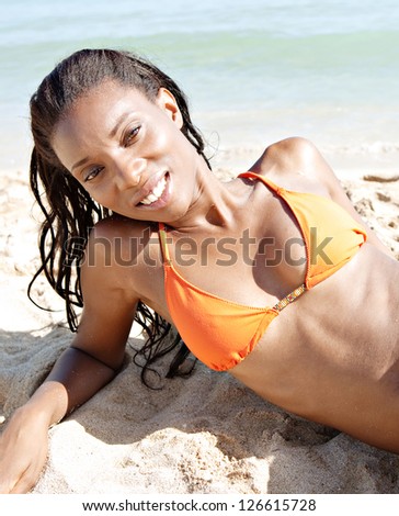 Portrait of a sexy black woman laying down on the shore of a white sand beach, relaxing and smiling at the camera with the sea behind.
