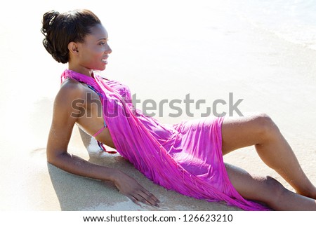 Side view beauty portrait of an attractive black woman wearing a bright pink sarong and relaxing on the beach shore, contemplating the sea.