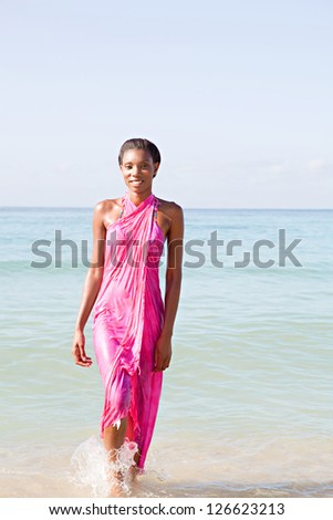 Happy black woman on a beach, walking out of the sea wearing a bright pink sarong around her body while on vacations.
