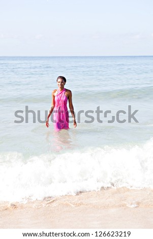 Happy black woman on a beach, walking out of the sea wearing a bright pink sarong around her body while on vacations.