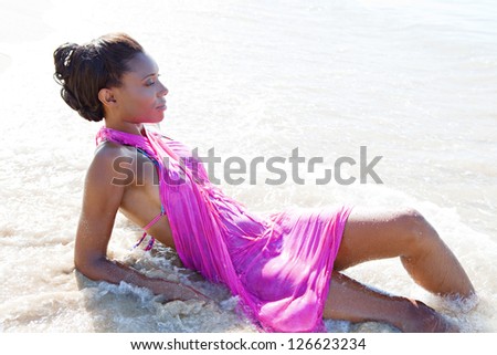 Side view beauty portrait of an attractive black woman wearing a bright pink sarong and relaxing on the beach shore, contemplating the sea.