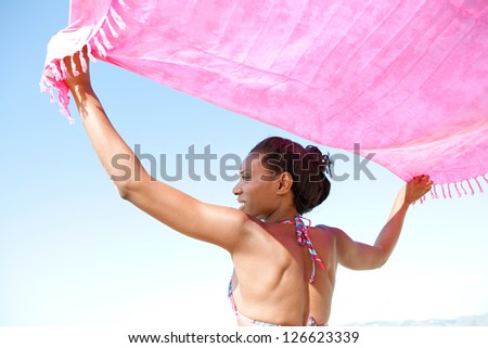 Back view of an attractive african american woman holding an bright pink sarong in the air with her arms raised against a clean blue sky background.