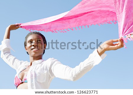 Aspirational beauty portrait of an african american woman on the beach, holding pink floating fabric above her head against a deep blue sky.