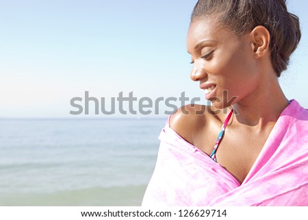 Close up profile portrait of an attractive black woman covering her shoulders with a pink sarong on the beach, smiling against a blue sky.