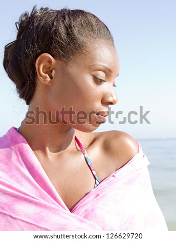Close up profile portrait of an attractive black woman covering her shoulders with a pink sarong on the beach, being thoughtful against a blue sky.