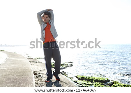 Full body view of a sports man stretching his arms after exercising by the sea, next to the water, against a blue sky background.