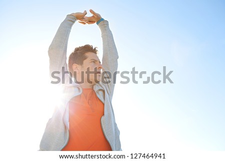 Close up portrait of a sports man stretching his arms up in the air against a sunny blue sky with sun rays filtering through his body.