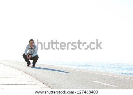 Sports man crouching down on running track by the sea, with a blue sky in the background on a sunny day, being thoughtful.