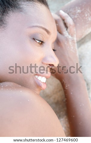Profile close up beauty portrait of an attractive black woman with perfect skin, laying down on a white sand beach, relaxing and smiling.
