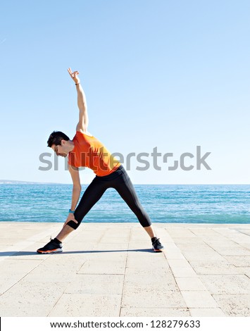 Wide view of a sports man body figure stretching on a track along the sea with the blue sky in the background and open space around him.