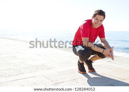 Portrait of a sports man crouching down on a track by the sea on a sunny day, smiling against a blue sky.