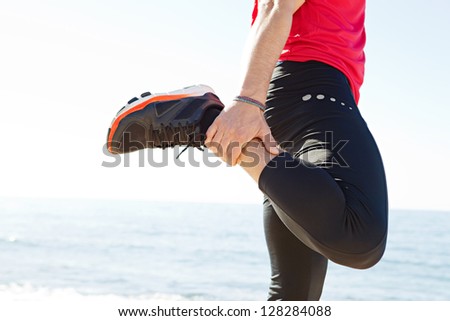 Middle section of a sports man body stretching his leg back while standing by the sea on a sunny day, against a blue sky.