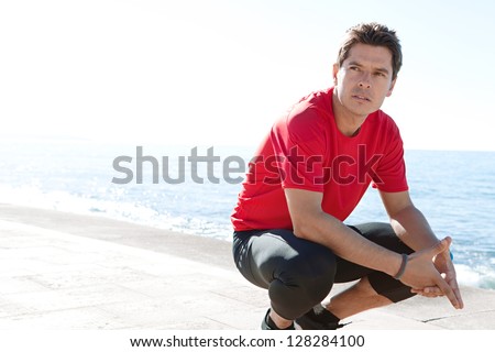 Portrait of a sports man crouching down on a track by the sea on a sunny day, being thoughtful against a blue sky.