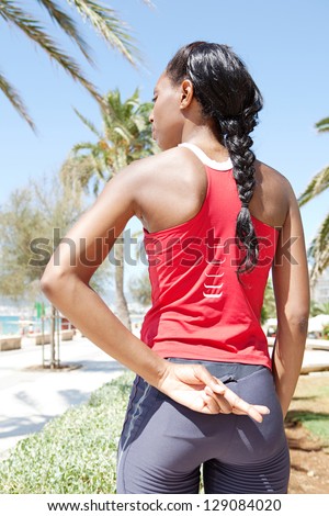 Rear view of a black woman standing against a blue sky after exercising, keeping her fingers crossed behind her back, in a coastal city.