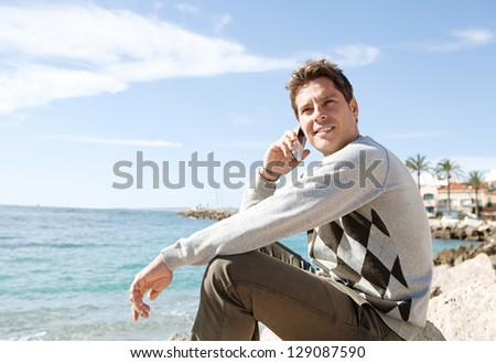 Side view of a professional man sitting on a rock on the beach having a phone conversation on his \