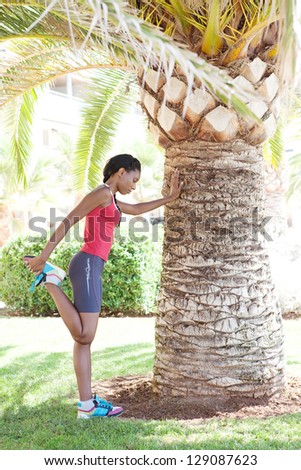 Side full body view of a black woman stretching her leg after exercising in the city, leaning on a palm tree trunk during a sunny day.