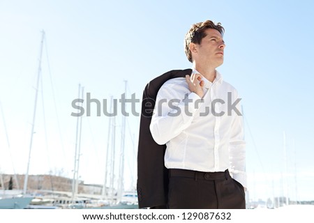 Aspirational successful businessman standing by luxury boats and yachts in a coastal city against a deep blue sky on a sunny day.