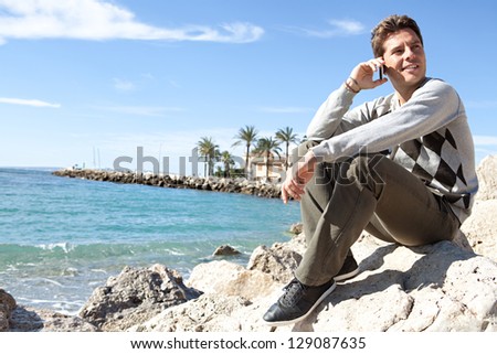Side view of a professional man sitting on a rock on the beach having a phone conversation on his \