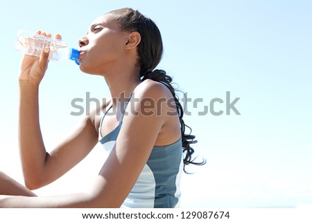 Attractive african american woman drinking mineral water from a bottle after doing sport against a blue sky background.