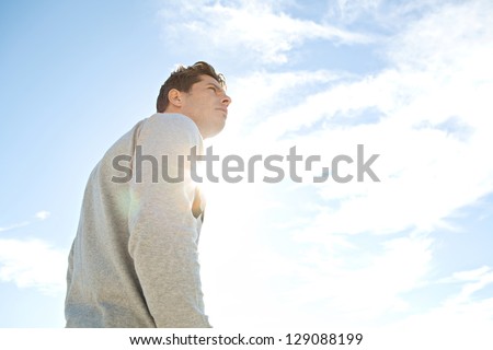 Side portrait low angle view of a man standing and looking ahead against a blue sky with sun rays filtering through his chest.