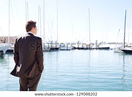 Rear view of a powerful businessman standing by expensive sailing boats and yachts in a coastal city, contemplating the views.