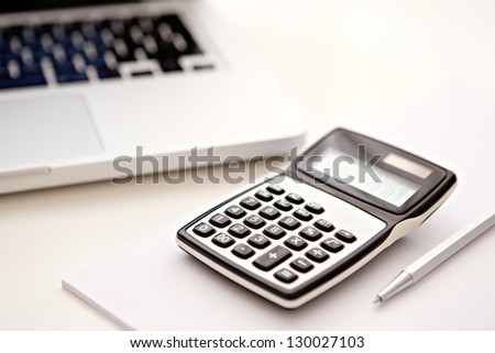 Close up detail view of a white work desk with a laptop computer and a calculator with numbers on the screen.