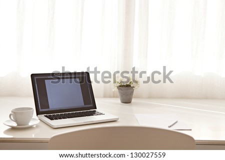 Wide view of a work desk interior with a laptop computer and white curtains on a sunny day.