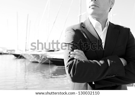 Middle section of a smart businessman wearing a suit and standing by a luxury yachts marine with his arms crossed. Black and white image.