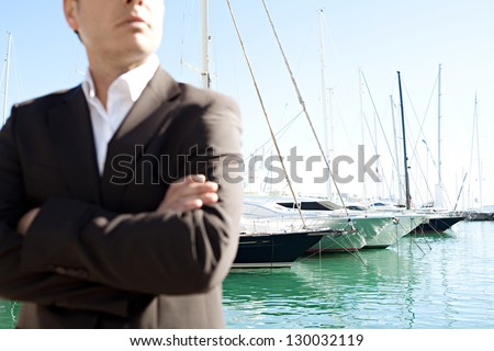 Close up middle section of a businessman standing by a luxurious yachts marine with his arms crossed against a deep blue sky and sea.