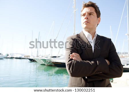 Close up portrait of an elegant businessman standing by a luxury yachts marine with his arms crossed against a blue sky, being thoughtful.