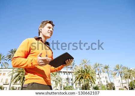 Side portrait of a business man using a digital tablet device with headphones while standing in a classic city against a deep blue sky.