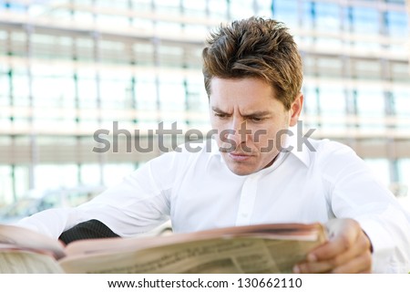 Close up portrait of a worried professional business man reading a financial newspaper while sitting near a modern office building in the city.