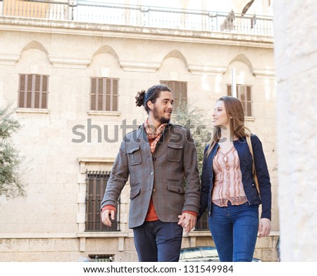 Trendy tourist couple walking passed classic architecture buildings in a destination city while on vacation, smiling and holding hands.