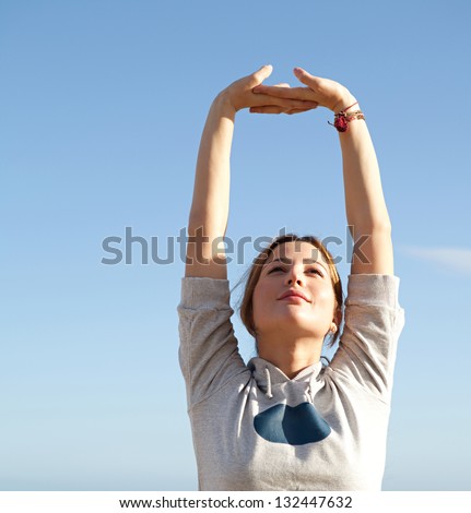 Young sports woman stretching her arms up with interlinked fingers while exercising on a beach with a blue sky and the sea in the background.