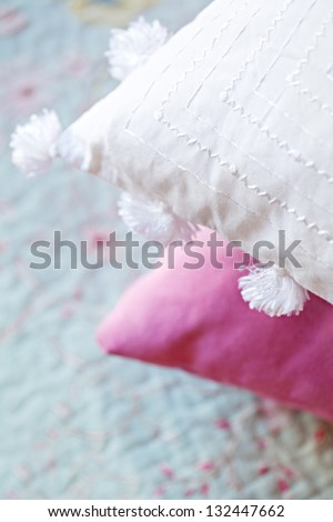 Close up detail view of an embroidered white cushion and a pink cushion laying on a soft blue bed cover in a home.