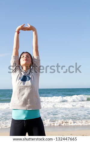 Young sports woman stretching her arms up with interlinked fingers while exercising on a beach with a blue sky and the sea in the background.