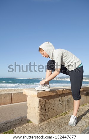 Sporty woman bending over to tie her shoe laces while exercising by a golden beach with an intense blue sky.