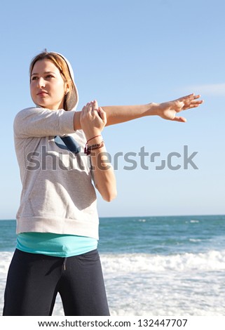 Healthy young woman exercising and stretching while standing on a beach with an intense blue sky and the sea in the background.